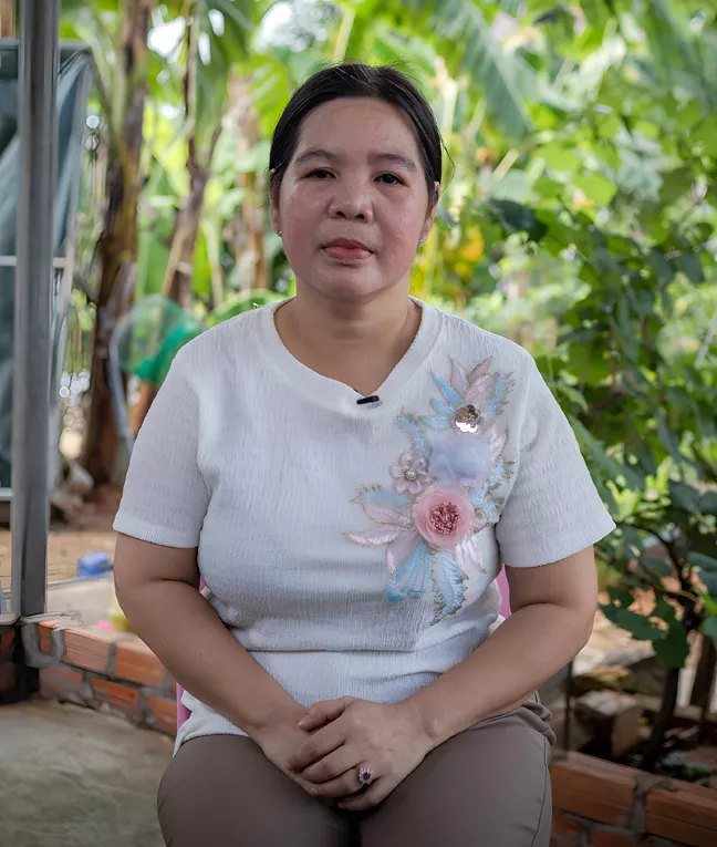 En medelålders kvinna sitter i en trädgård. Hon har händerna i knäet och tittar in i kameran.  